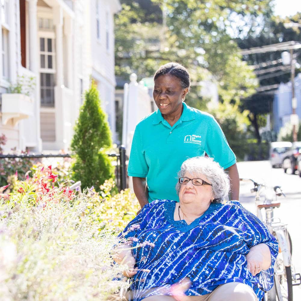 Female caregiver pushing a senior female in a wheelchair outside next to a bush in front of a home