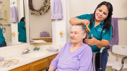 A female caregiver wearing a mask stands behind male patient while brushing his hair.