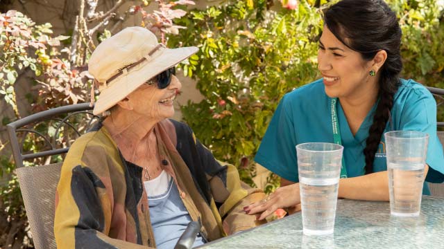 A female caregiver sits across from an elderly female patient outside with glasses of water. They both smile.