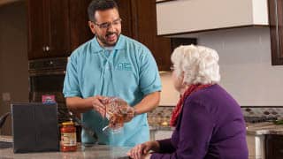 A male caregiver holds meal ingredients while talking and smiling with a female patient.