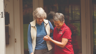 A female caregiver in red helps an elderly woman walk down stairs. They both smile.