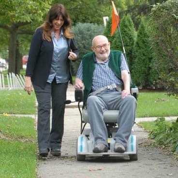 A female caregiver walks on the sidewalk with male senior in a wheelchair. 